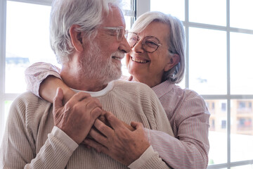 Smiling senior family couple of Caucasian people hugging in front the window, elderly man and woman...