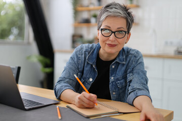 Senior woman works on a laptop and writes a reminder in a notebook.