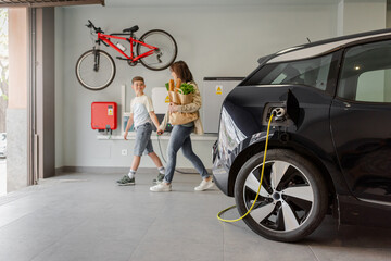 Electric vehicle charging station in private home with happy mother and son walking alongside, blurred family leaving the house, with a bicycle hanging on the wall. Horizontal. - obrazy, fototapety, plakaty