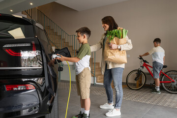 Smiling mother and son charging electric car at home. Boy helps woman to plug a charger yellow cable from the charging home station. Sustainable Alternative Healthy Lifestyle. Horizontal, copy-space