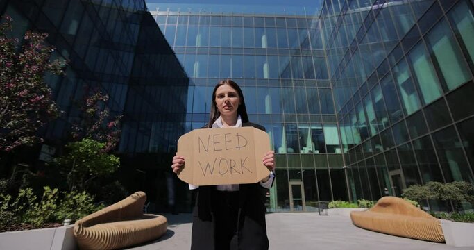 Portrait of woman in a black suit holding Need Work sign searching job standing near office centre. Jobless people concept. Global unemployment.