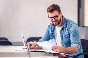 Young businessman sitting at the desk in the office, looking at papers and writing down.