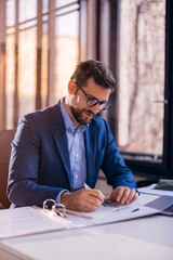 A young businessman is sitting at a desk in the office, working on a laptop and writing down ideas...