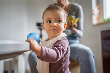 One year old girl playing with blue ball while her mother feeding her at home, early child...
