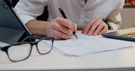 Close up. Man in stylish clothes sitting at kitchen table and working with important documents using information on laptop.