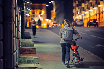 Woman with backpack walk alone on night city street with electric scooter. Girl rolls scooter with...