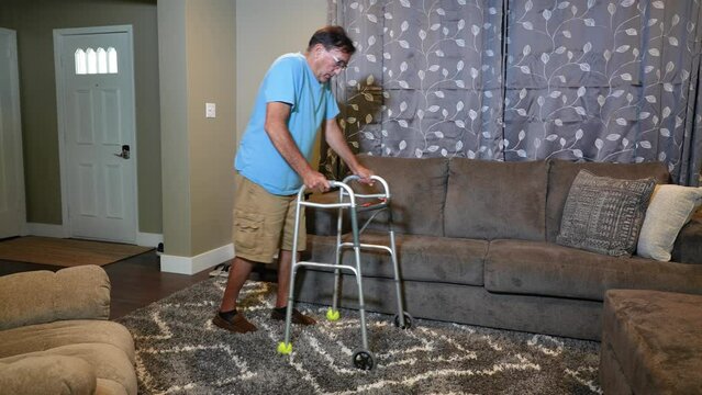 A wide shot of a man in his living room using a walker to move to the sofa and sit down.  	