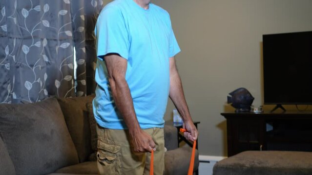 A tilt up medium shot view of a man using a rubber exercise band in his living room at home.  	