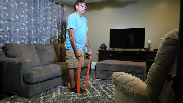 A tilt up wide shot view of a man using a rubber exercise band in his living room at home.  	