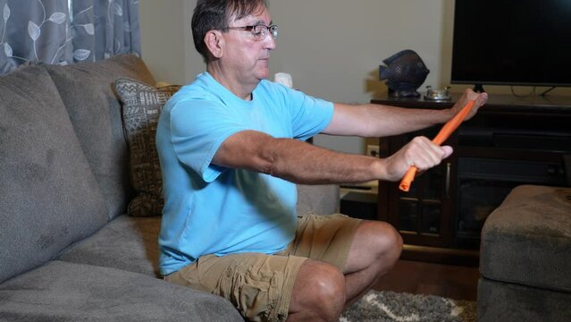 A profile view of a man using an orange rubber exercise band while sitting on his couch in the living room.  	