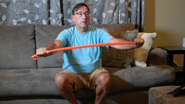 A front view of a man using an orange rubber exercise band while sitting on his sofa in the living room.  	