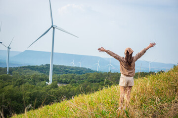 Woman wearing a cute dress stands spread arms on a meadow with wind turbine and mountains in the...
