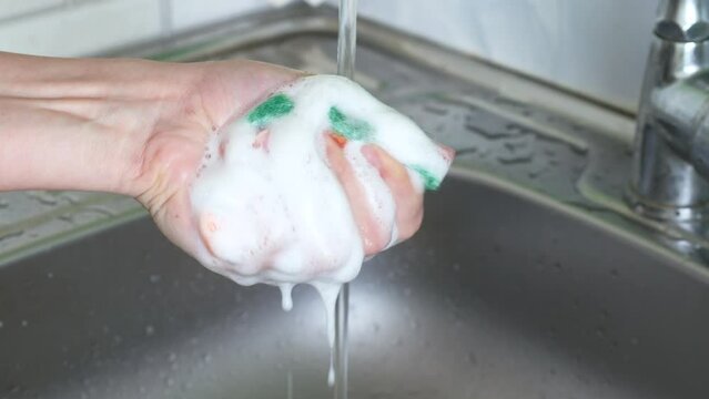 Foam from dishwashing liquid on kitchen sponge in the hand close up. Woman hands holding a cleaning sponge with dish wash gel. Household work concept