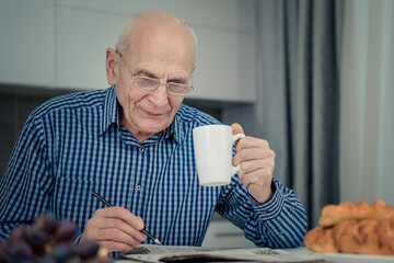 Happy senior man with pen in hand wearing eyeglasses solving crossword and drinking coffee sitting at kitchen table indoor. - obrazy, fototapety, plakaty