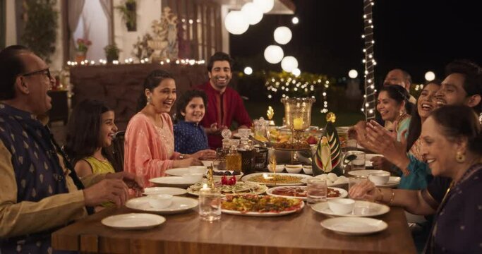 Happy Extended Indian Family Dining Together Outdoors in Fancy House Garden at Night. Family Members Laughing and Sharing Funny Stories and Memories While Eating Delicious Food. Slow Motion Arc