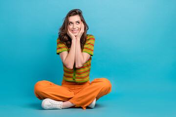 Full size photo of minded woman dressed orange pants colorful cardigan look empty space arms cheekbones isolated on blue color background