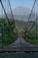 Fototapeta na wymiar Wooden suspension bridge over the river with beautiful mountain views. Rural scene of Indonesian countryside - Magelang, Indonesia