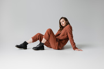full length of trendy model with brunette and wavy hair sitting in trendy and oversize suit with terracotta blazer, pants and black boots, looking at camera on grey background, stylish young woman - obrazy, fototapety, plakaty