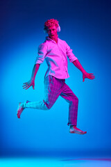 Full-length portrait of mature man listening to music in headphones and jumping against blue studio background in pink neon light. Concept of human emotions, lifestyle, youth, facial expression