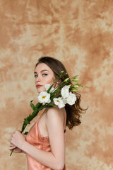 Obraz na płótnie Canvas young woman with brunette hair posing in pink and silk slip dress and holding white eustoma flowers on mottled beige background, sensuality, sophistication, elegance, looking at camera