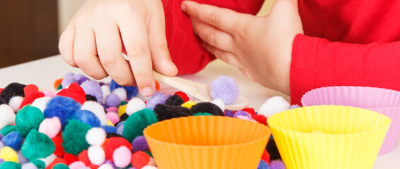 Preschooler playing with small pompoms, wooden spoon and cups. Development of kids motor skills, coordination and logical thinking