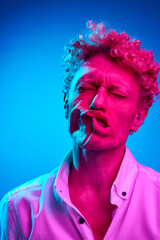 Funny image of man with curly hair posing with blowing wind on face against blue studio background in pink neon light. Concept of human emotions, lifestyle, youth, facial expression