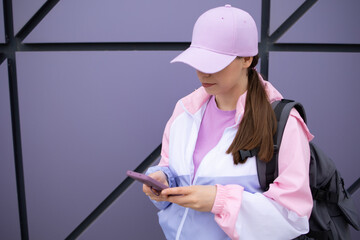 Woman no face, using a phone in a purple silicone case with ring. In a stylish jacket in pink, lilac, white colors and a baseball cap on a plain wall. Copy space. Mockup