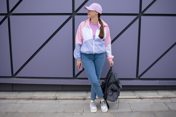 Woman in a pink jacket, a lilac baseball cap, jeans, backpack standing in full growth near the wall. Mockup. Copy space. There is no face. Stylish fashion, sporty clothing style, 90-80 style fashion