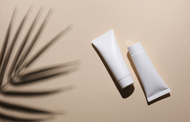 two white cream tubes on beige background with palm leaf shadow. Creative beauty layout.