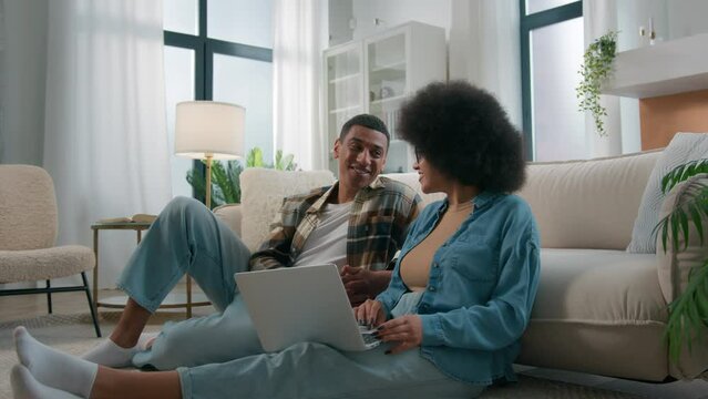 African American couple on floor at home using online laptop computer browsing shopping together pay bills choose products order delivery do high five gesture teamwork happy family in own apartment