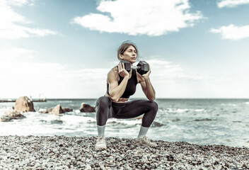 Strong athletic woman exercising with heavy dumbbell on the beach during the day with blue sky and clouds. Functional outdoor training