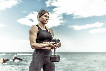 Strong athletic woman exercising with heavy dumbbell on the beach during the day with blue sky and clouds. Functional outdoor training