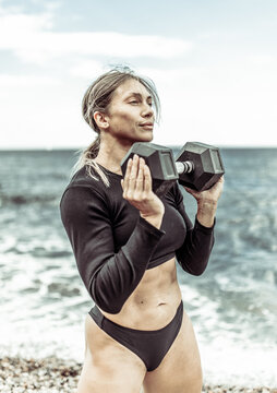 Strong athletic woman exercising with heavy dumbbell on the beach during the day with blue sky and clouds. Functional outdoor training