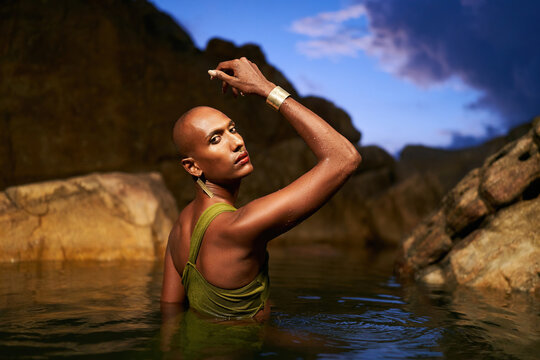 Non-binary person of color poses in water, shows jewelry inside scenic creek at night. Lgbtq ethnic graceful fashion model wears brass rings with gems, nose-ring, earrings, bracelets immersed in pond.
