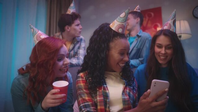 An African American girl shows the phone to her friends, they are emotionally looking at photos, videos or talking on a video call. A group of friends in party hats in a room decorated for a party.