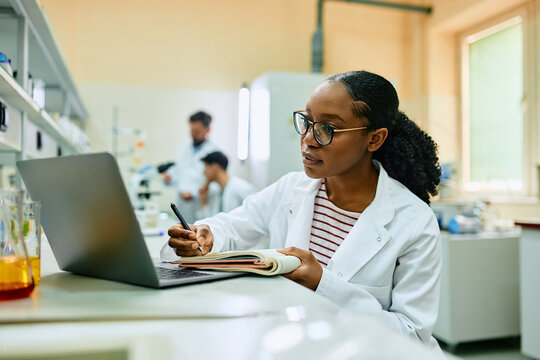 Black female biochemist writes research data in notebook while working on laptop in laboratory.