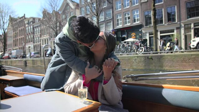 Two LGBT girls share a kiss and embrace on a sunny day while sailing through picturesque canals in Amsterdam. Medieval style houses float past in the city's enchanting ambiance.