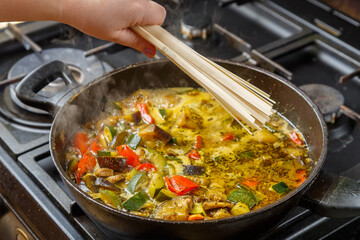 A woman's hand puts udon in a frying pan with stewed vegetables in spices.