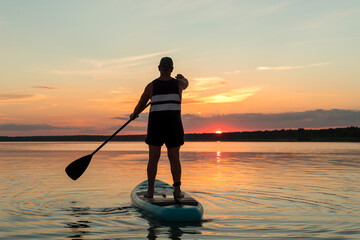 A man in shorts stands on a SUP board with an oar at sunset in a lake against the backdrop of a sunset sky.