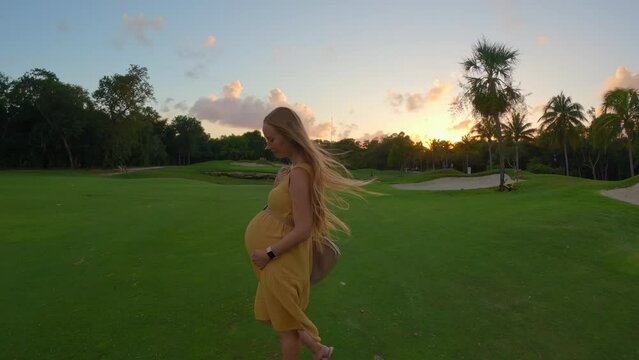 In this mesmerizing slow-motion stock video, a pregnant woman walks gracefully on a vibrant green field, illuminated by the enchanting hues of a beautiful sunset. The serene atmosphere and the woman's