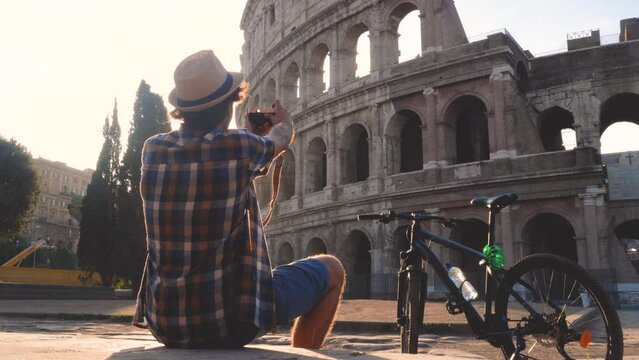 Happy young man tourist wearing shirt and hat with bike sitting and taking pictures with vintage camera at colosseum in Rome, Italy at sunrise.