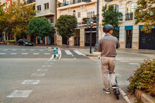 A guy stands on a pedestrian crossing and waits for a green light. Back view of young man on vacation having fun driving electric scooter through the city. High quality photo