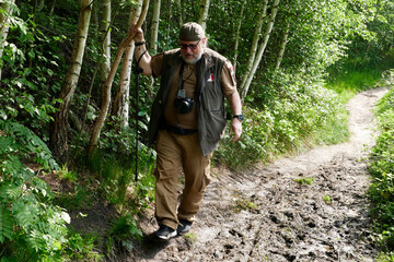 Active senior man of 70 with camera and walking stick tries to keep his shoes out of the mud and...