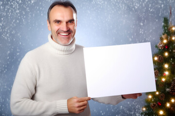 Happy man holding white sheet of paper in front of christmas tree