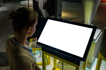 Woman looking at blank white display of interactive kiosk at exhibition or museum with futuristic...