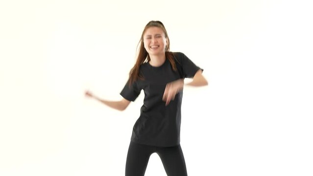 Pretty woman dancing modern dance on white wall background. She has long hair and wearing black leggings and black T-shirt. Joyful girl enjoying her favorite music.