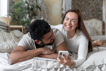 Multiethnic American couple using smartphone lying on bed together