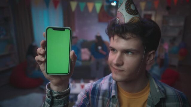 A guy shows a smartphone with a green screen, smiles and nods his head. Close up of a guy with a smartphone on a blurred background of a party, in a room decorated with a disco ball and paper flags.