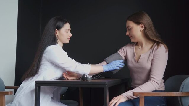 A young female cardiologist in a white coat uses a tonometer to measure the blood pressure of a young woman during an examination in a clinic office