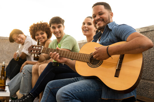 A group of 5 young interracial millennials are sitting on a one-story terrace drinking beer and having fun.The African boy in the foreground plays a classical guitar.Concept of interracial music group